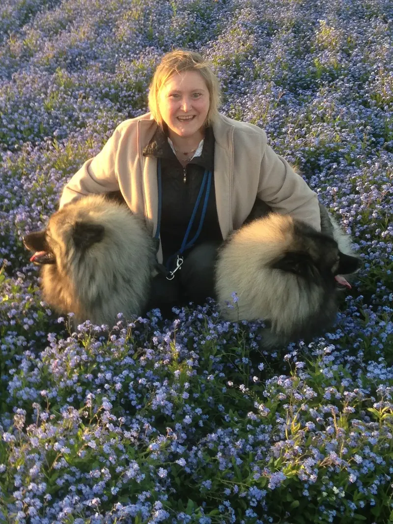 Renate kniet mit zwei Wolfsspitzen in einer blühenden Wiese voller Vergissmeinnicht